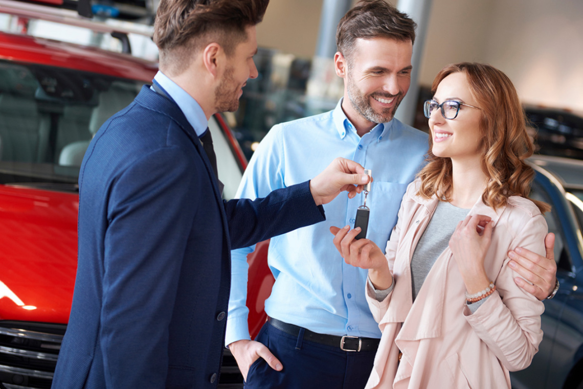 Car dealer handing keys to a couple, standing in front of a red SUV