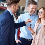 Car dealer handing keys to a couple, standing in front of a red SUV